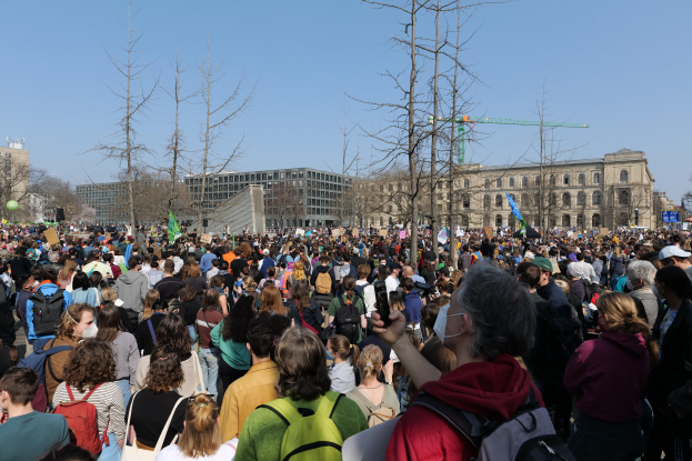 Eine große Menge Menschen steht vor einem Gebäude mit Fenstern, Bäumen und einem klaren Himmel, viele halten Schilder und tragen Taschen, was auf eine Klimaprotestaktion hinweist.