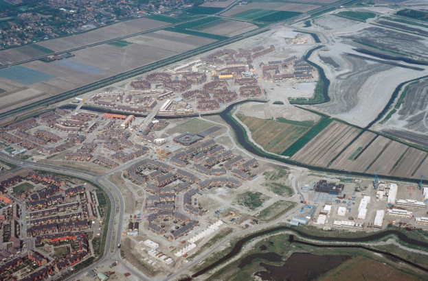 Luftaufnahme einer Stadt mit dichtem Gebäudebestand, Straßen, Grünflächen, Bäumen und einem zentralen Reservoir umgeben von städtischer Infrastruktur.