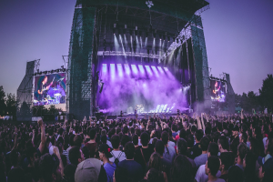 Eine große Menschenmenge bei einem Musikfestival schaut Künstlern auf einer hell erleuchteten Bühne mit Instrumenten und Bildschirmen zu, die vor einem Gebäude, Bäumen und einem klaren blauen Himmel aufgebaut ist.