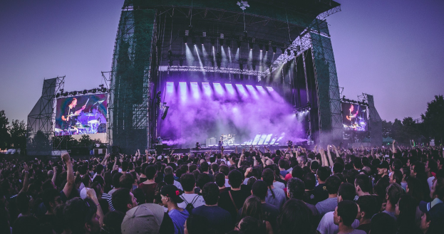 Eine große Menschenmenge bei einem Musikfestival schaut Künstlern auf einer hell erleuchteten Bühne mit Instrumenten und Bildschirmen zu, die vor einem Gebäude, Bäumen und einem klaren blauen Himmel aufgebaut ist.
