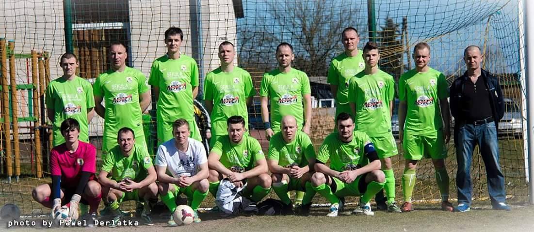 Männergruppe in grünen T-Shirts, die auf einem Fußballfeld mit Ball und Torpfosten stehen oder knien, umgeben von Bäumen, Gebäuden und Fahrzeugen.