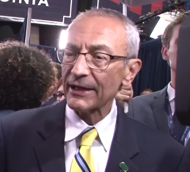 Ein Mann im Anzug mit Brille spricht zu Reportern über ein Mikrofon, umgeben von einer Menge, mit beleuchtetem Hintergrund und einer sichtbaren Tafel.