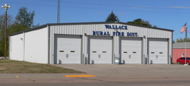 Feuerwache des Wallace Rural Fire District mit einer Straße darunter, Gras links, einem Lichtmast rechts, Bäumen im Hintergrund, einem zentralen Turm und einer Flagge, die über einem sichtbaren Himmel weht.