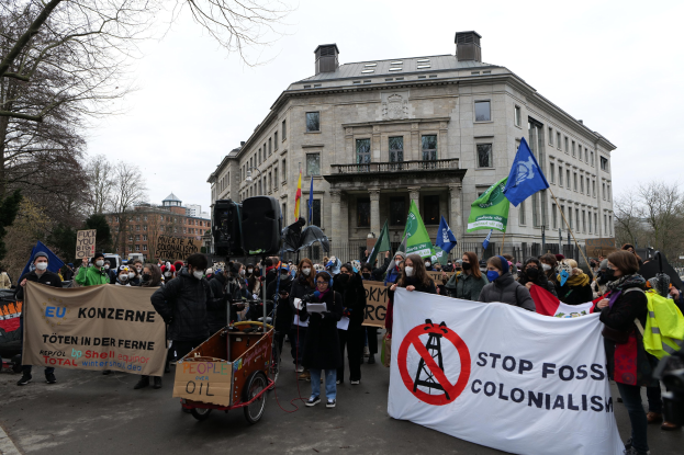 Eine große Gruppe von Menschen marschiert bei einer Demonstration gegen fossile Brennstoffe, trägt Transparente und Fahnen, mit einem Fahrzeug im Vordergrund und Gebäuden, Bäumen und einem klaren blauen Himmel im Hintergrund.