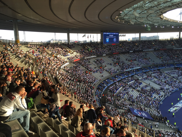 Großes Publikum in einem Stadion bei einem Fussballspiel mit einer Bühne, Fahnen, Masten, einem Bildschirm und der Allianz Arena in München, Deutschland im Hintergrund.