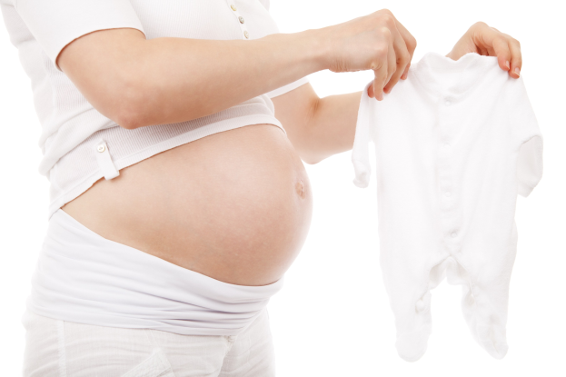 Eine schwangere Frau in einem weißen Kleid hält ein weißes Baby-Bodysuit und strahlt glücklich.