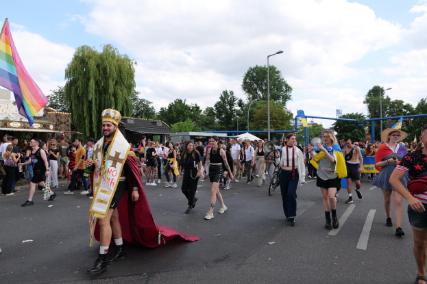 Eine Gruppe von Menschen, die bei der Pride Parade 2018 marschieren und eine Regenbogenflagge sowie Musikinstrumente tragen, mit Laternenmasten, Bäumen, Hütten und einem bewölkten Himmel im Hintergrund.