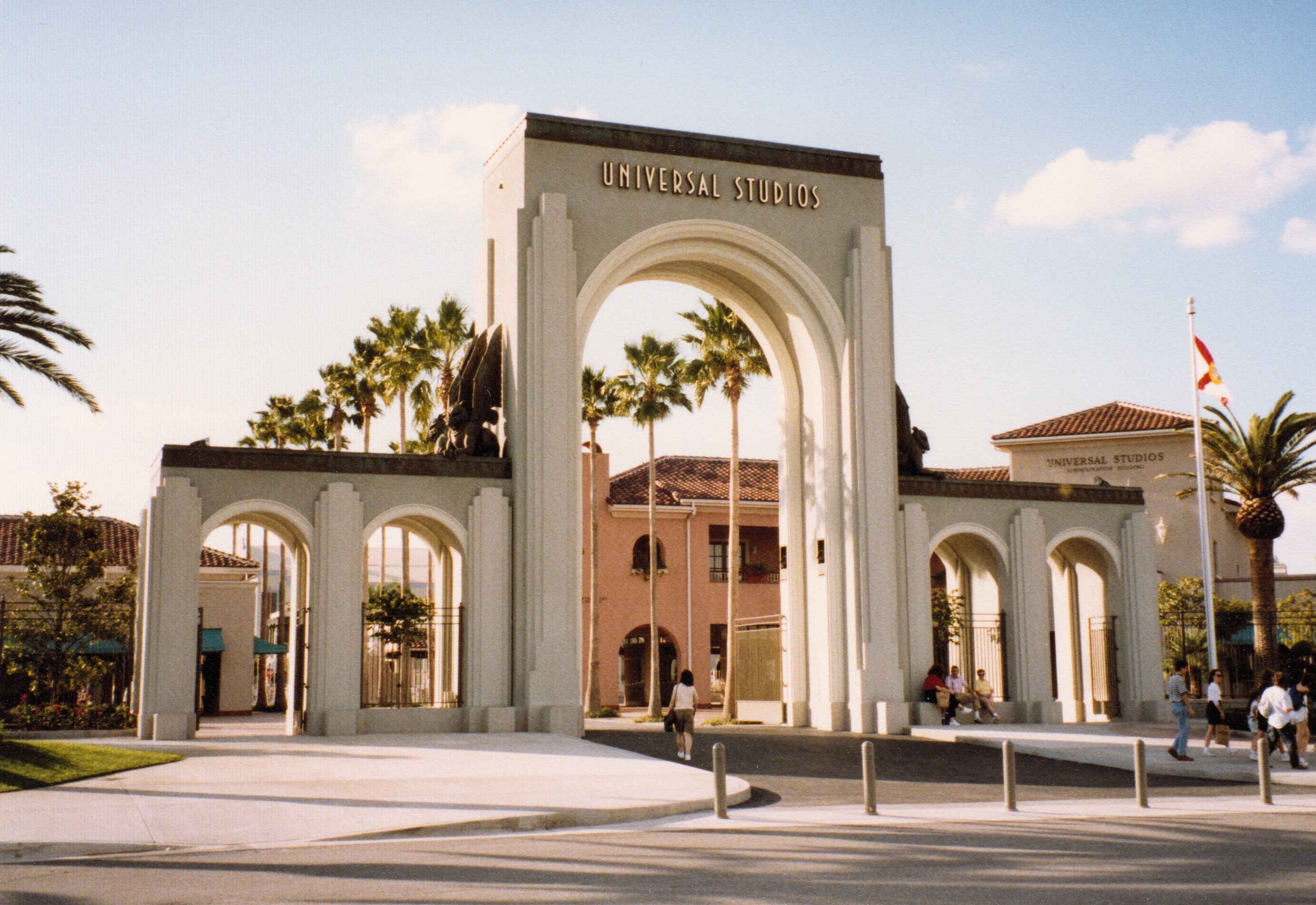 Eingang zu Universal Studios Hollywood mit einem Bogen, einer Statue, Menschen, Bäumen, Gebäuden, einem Fahnenmast mit einer Fahne und einem bewölkten Himmel.