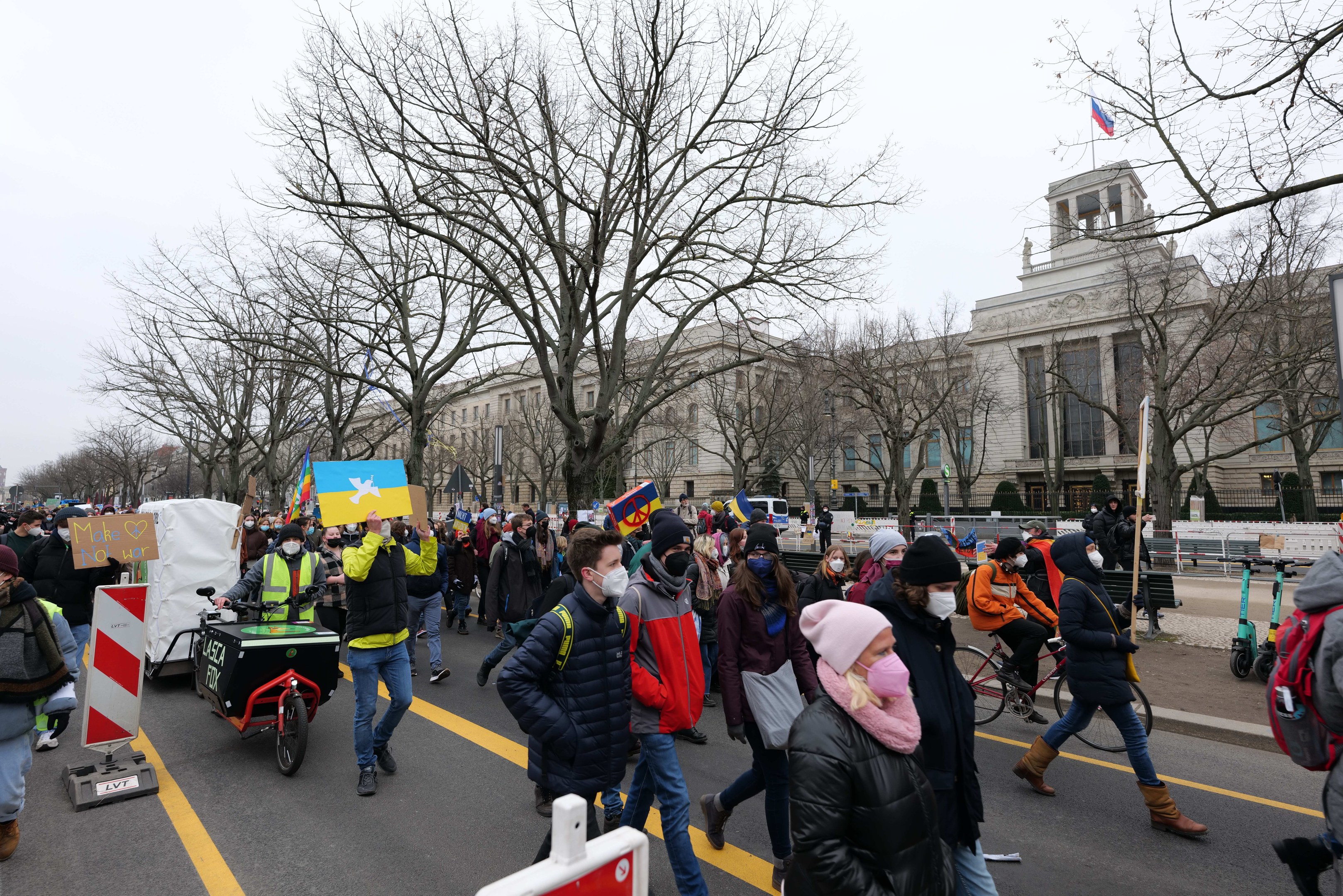 Eine große Gruppe von Menschen marschiert auf einer Stadtstraße in Washington, D.C., mit Schildern und Bannern, während einige Fahrrad fahren und Bäume und ein klarer Himmel im Hintergrund zu sehen sind.