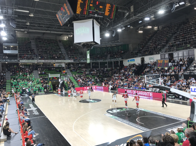 Basketballspiel in einer großen Arena mit Zuschauern, mit den Teams Asvel und Galatasaray, unter Stadionbeleuchtung und Anzeigetafeln.