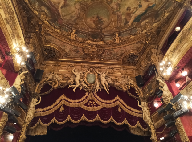 Innere eines Theaters mit roten Vorhängen, Kronleuchtern, Skulpturen, Lichtern und einer bemalten Decke, das eine Bühne mit ein paar Menschen am Boden zeigt.