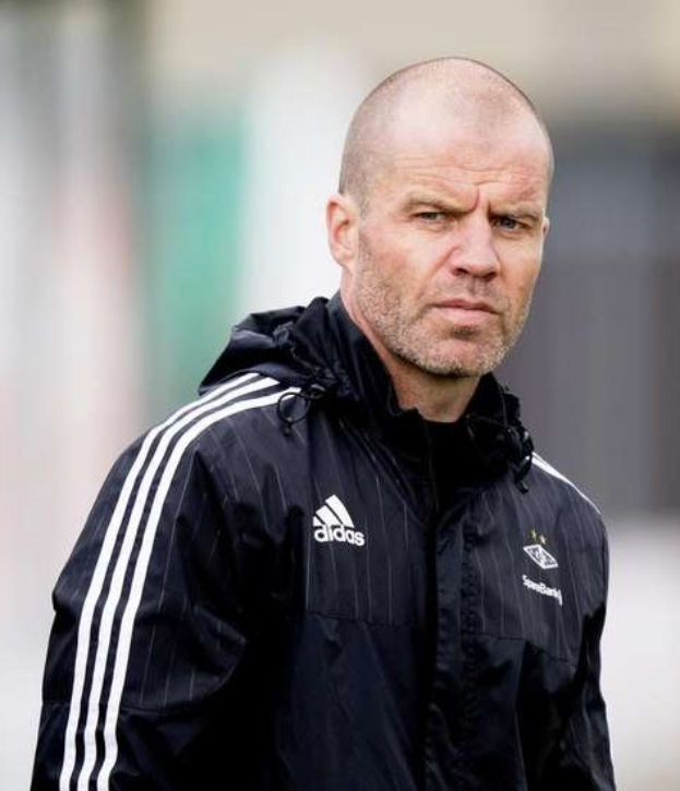 Glatzköpfiger Mann in einem blauen Jackett mit weißen Streifen und weißem Kragen, der auf einem Fußballfeld steht und die Hände verschränkt hält.