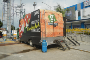 Lkw vor einem Gebäude mit einer Tafel, die "Teilen Sie eine positiv überladene Erfahrung" lautet, mit Stufen, einem Zylinder auf dem Boden, Geländern, Strom- und Lichtmasten, Bäumen und einem bewölkten Himmel.