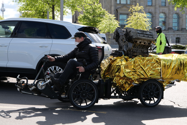 Ein Mann im Rollstuhl mit einem großen Motor am Rücken, umgeben von Fahrzeugen auf einer Straße, mit Bäumen, Gebäuden, Polen und einem klaren blauen Himmel im Hintergrund.