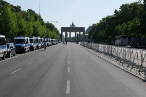 Lange Reihe von Polizeiwagen auf der Seite einer Straße vor dem Brandenburg-Tor in Berlin, Deutschland, mit Fahrradfahrern und Fußgängern, Barrieren, Bäumen, einem prunkvollen Tor mit Statuen und Himmel.