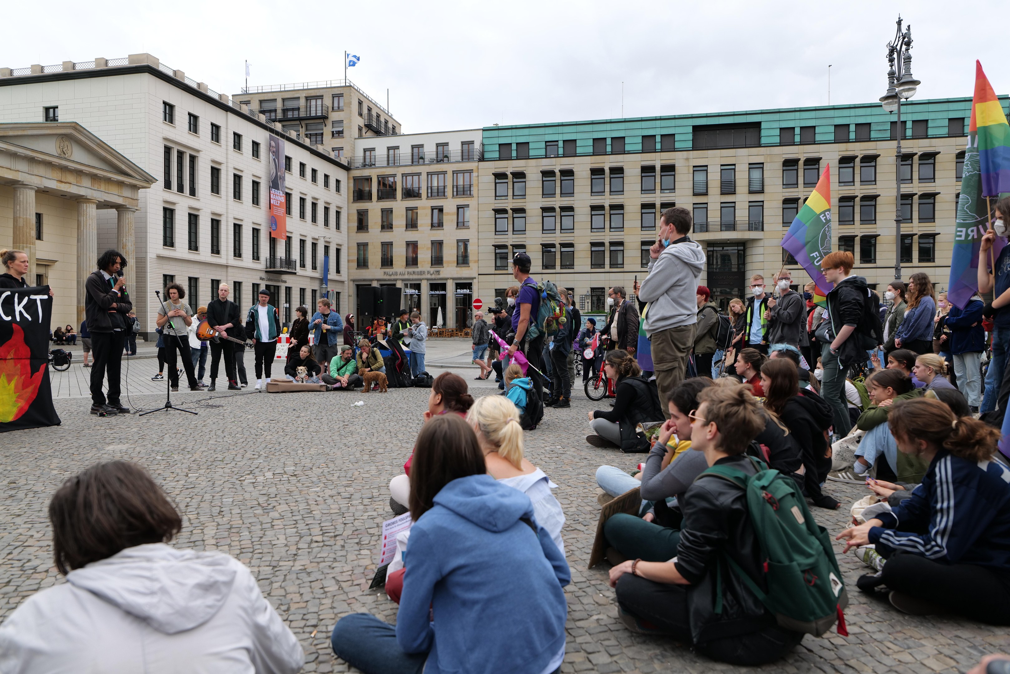 Eine Gruppe von Menschen auf dem Boden sitzend, die einer Menge mit Fahnen, Schildern und einem Mikrofon auf einem Ständer gegenüberstehen, mit einer Statue, Gebäuden und einem bewölkten Himmel im Hintergrund während einer anti-schwulen Demonstration in Berlin, Deutschland.
