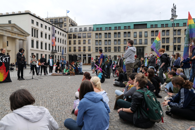 Eine Gruppe von Menschen auf dem Boden sitzend, die einer Menge mit Fahnen, Schildern und einem Mikrofon auf einem Ständer gegenüberstehen, mit einer Statue, Gebäuden und einem bewölkten Himmel im Hintergrund während einer anti-schwulen Demonstration in Berlin, Deutschland.