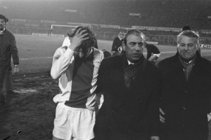 Eine Gruppe von Männern steht zusammen auf einem Fußballfeld, sieht besorgt aus, mit einem Stadion im Hintergrund, dargestellt in Schwarz-Weiß.