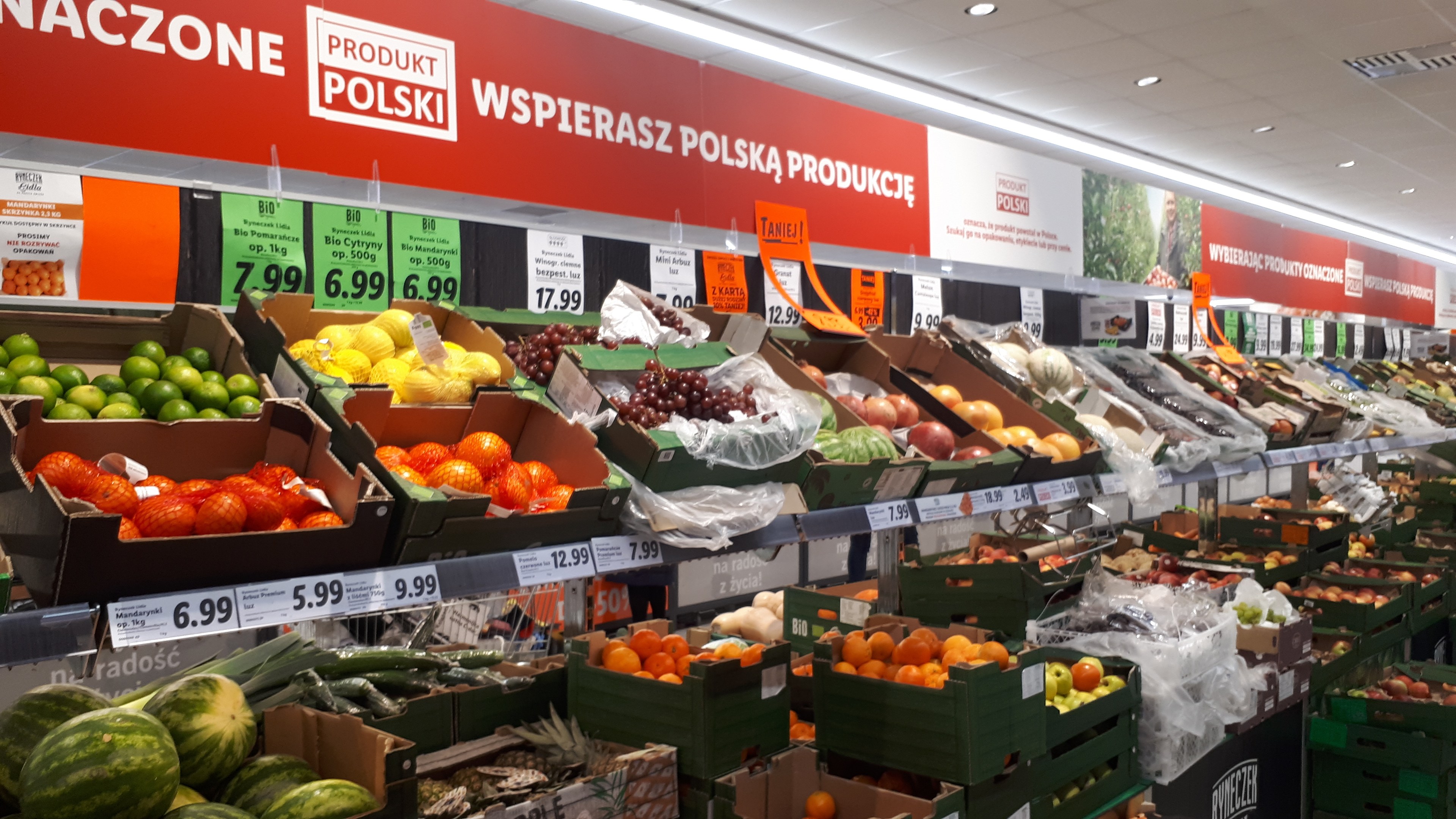 Ein Geschäft mit verschiedenen Obst- und Gemüsesorten in Kartonschachteln mit Preisschildern, Informationsschilder an den Wänden und Deckenbeleuchtung.