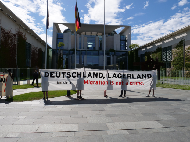 Eine Gruppe von Menschen hält ein Transparent mit der Aufschrift "Deutschland, Lagerland Migration ist kein Verbrechen" vor einem eingezäunten Gebäude mit Bäumen und Flaggen unter einem bewölkten Himmel.