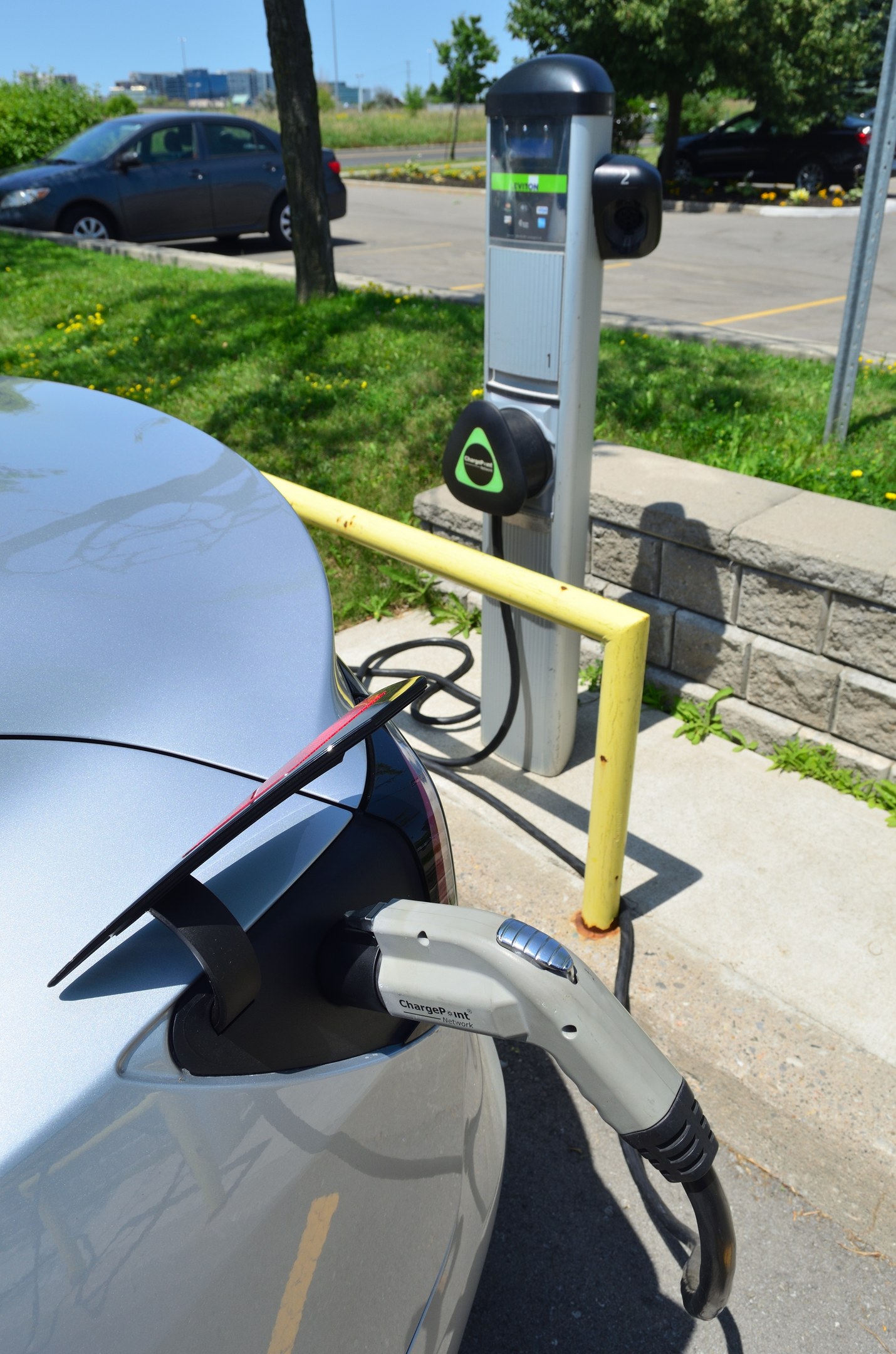 Ladestation für Elektroauto am Straßenrand mit Gras, Bäumen, Strommasten und Gebäuden im Hintergrund bei klarem blauem Himmel.