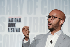 Ein Mann im Anzug und mit Brille, der auf einer Bühne spricht und ein Mikrofon trägt.