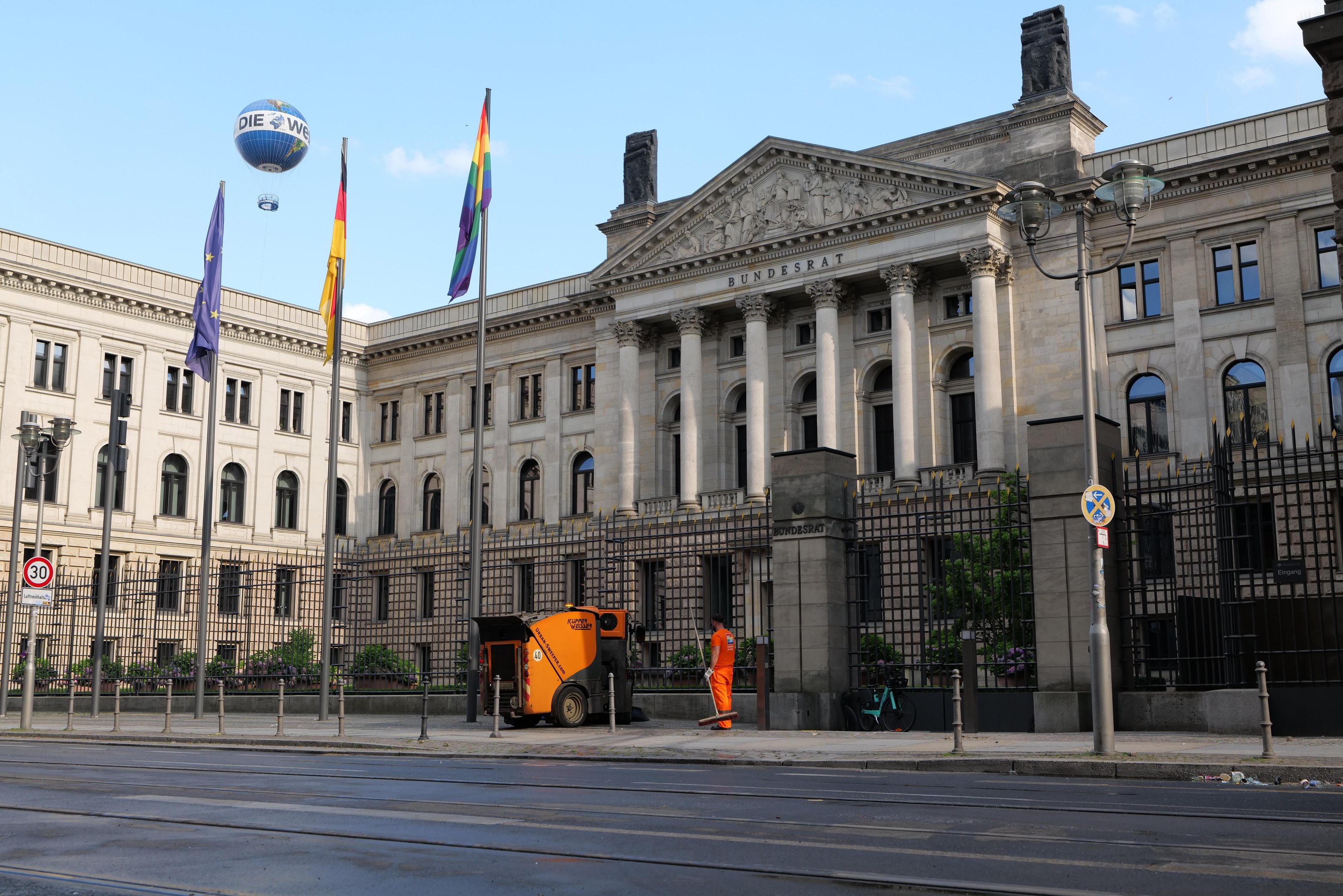 Großes modernes Regierungsgebäude mit vielen Fenstern, umgeben von Straßeninfrastruktur, Fußgängern und Bäumen, mit einem bewölkten Himmel im Hintergrund.