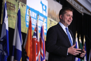 Ein Mann im Anzug spricht an einem Podium mit Mikrofonen, umgeben von Flaggen und einem Bildschirm, wahrscheinlich zu einer Regierungszahlung an die Milchindustrie.