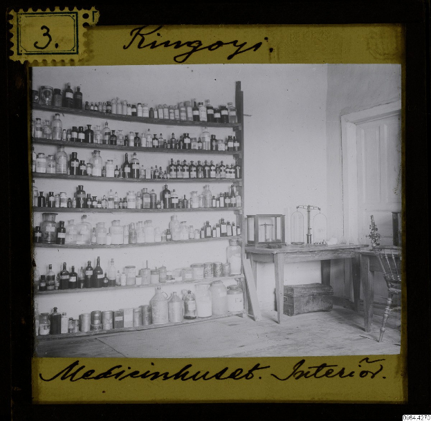 Schwarz-weiß-Foto einer alten Apotheke mit Regalen voller Flaschen, einem Tisch mit Gegenständen, einem Stuhl, einer Tür und einer Wand mit Text unten.