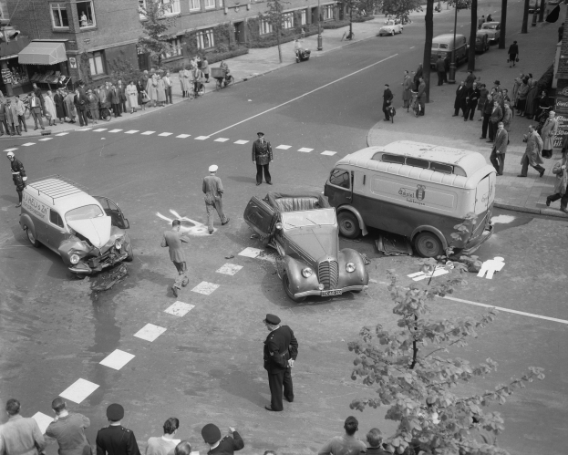 Eine Schwarz-Weiß-Szene eines Autounfalls an einer Straßenkreuzung mit mehreren Fahrzeugen, darunter ein Lieferwagen, einer Gruppe von Menschen drumherum und im Hintergrund Gebäude, Bäume, Masten und Texttafeln.