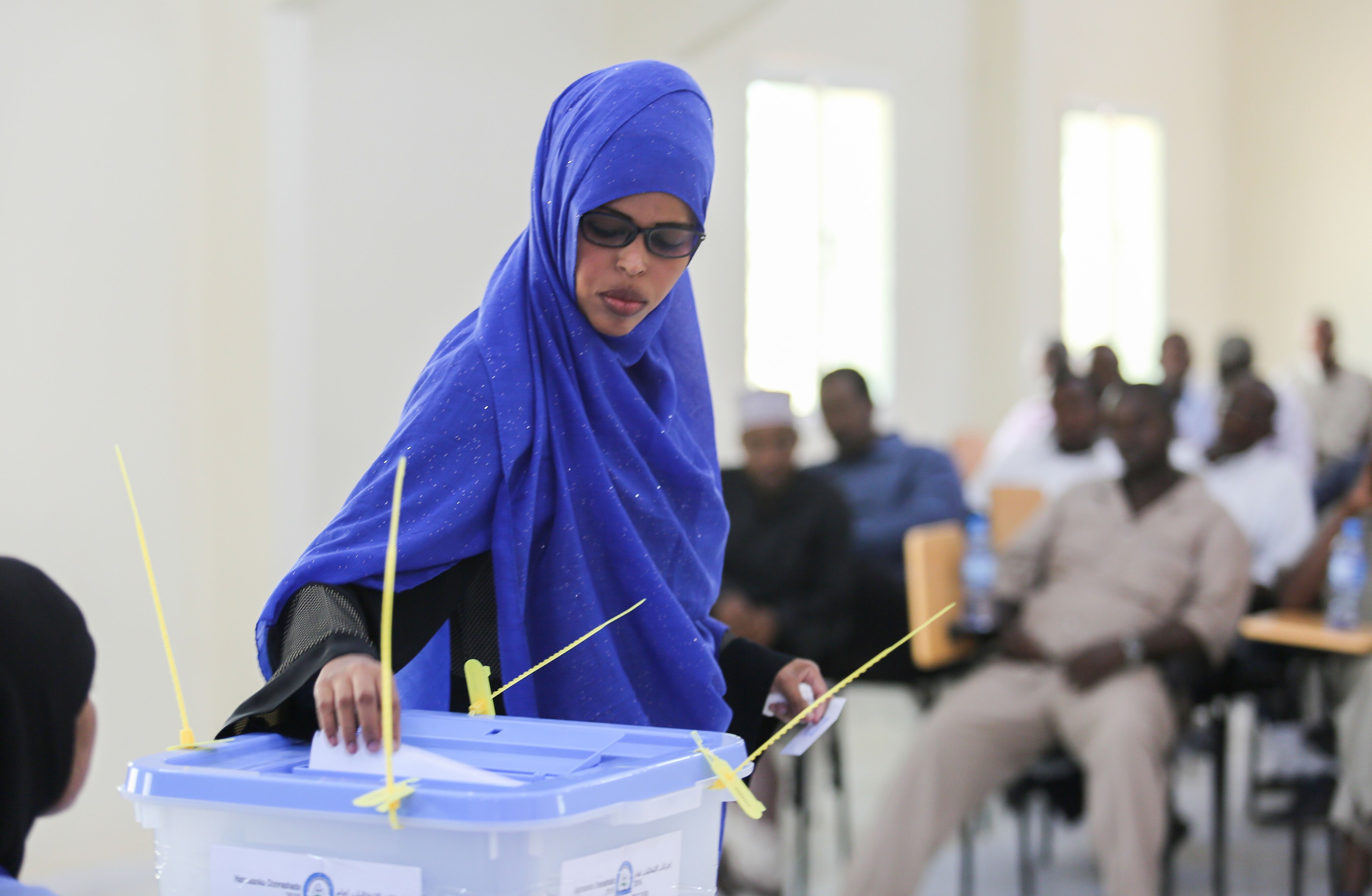 Eine Frau mit einem blauen Kopftuch gibt ihre Stimme in einer Wahllokal ab, während andere in der Nähe sitzen.