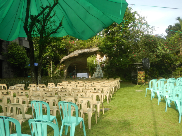 Eine Hochzeitszeremonie im Garten mit grünen Stühlen und Schirmen, umgeben von üppiger Vegetation, mit einem Gebäude und einer Scheune im Hintergrund und dem Himmel oben sichtbar.