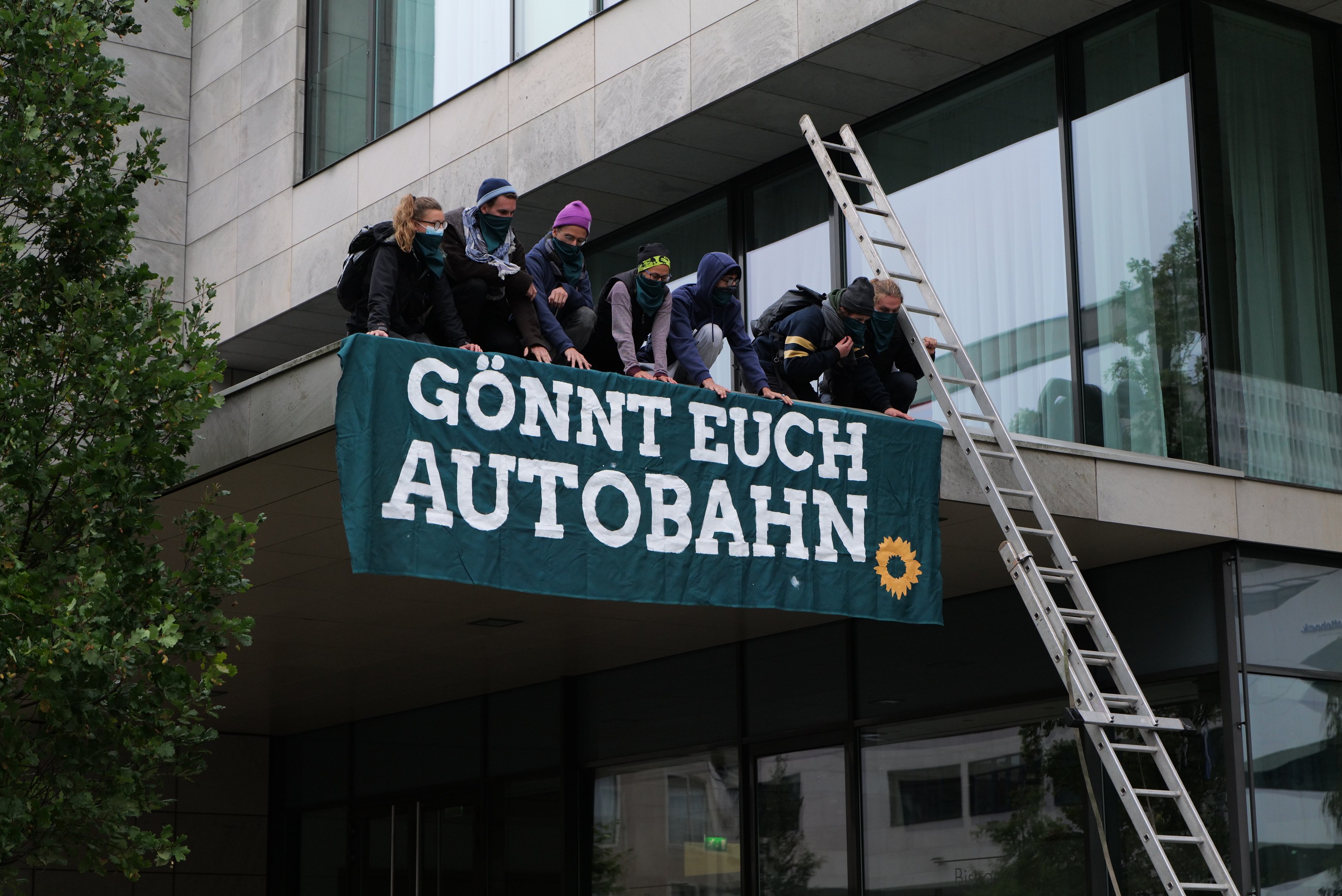 Gruppe von Menschen auf einem Gebäuredach mit einer Fahne, mit einem Baum links und Glasfenstern im Hintergrund, wahrscheinlich protestierend für die deutsche Regierung.