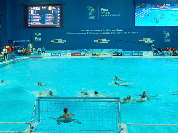 Wasserballspieler bei den 2016 FINA-Weltmeisterschaften in Kazan im Pool gegeneinander antretend, mit Zuschauern links und einer Anzeigetafel mit Event-Branding im Hintergrund.
