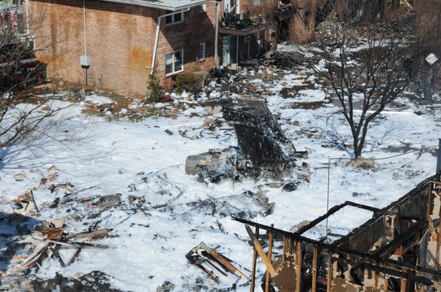 Verbrannte überreste eines durch Feuer zerstörten Hauses in einer schneebedeckten Wohngegend, mit Bäumen und Gebäuden im Hintergrund.