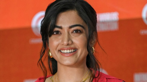 Eine Frau in einem roten Kleid mit einem eleganten Dutt und dezentem Make-up lächelt in die Kamera, trägt schwarze High Heels und hat einen hellgelben Banner mit der Aufschrift "Sonam Kapoors schönste Looks vom Cannes Film Festival" im Hintergrund.