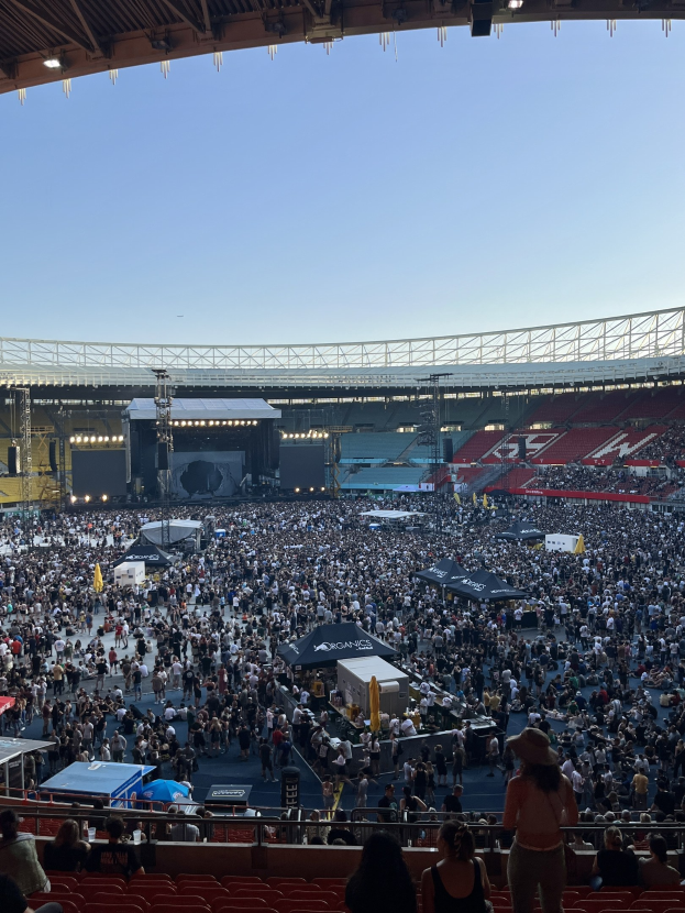 Eine große Menschenmenge sitzt und steht in einem Stadion mit Geländern, Zelten und Lichtern unter einer Decke mit Überkopfbeleuchtung, mit dem Himmel im Hintergrund.