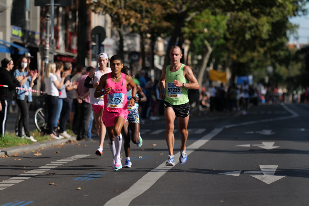Gruppe von Menschen, die bei einem Marathon auf einer Stadtstraße laufen, mit Zuschauern auf der linken Seite, unscharfen Hintergrund mit Gras, Bäumen, Gebäuden, Polen, Brettern und einem Fahrrad.