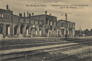 Schwarzes und weißes Foto einer Bahnstation in Korschen Ost, Deutschland, mit Bahnschienen, Gebäuden, Strommasten, Menschen, Bäumen und Himmel.