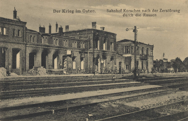 Schwarzes und weißes Foto einer Bahnstation in Korschen Ost, Deutschland, mit Bahnschienen, Gebäuden, Strommasten, Menschen, Bäumen und Himmel.