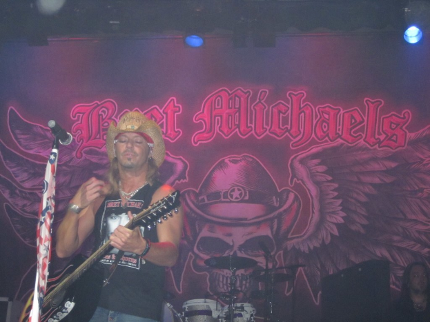 Musikalisches Konzert mit Scheinwerfern, einem Banner im Hintergrund, einer sitzenden Person rechts unten, einem stehenden Gitarristen links mit Mikrofon davor und Gegenständen in der Mitte unten.