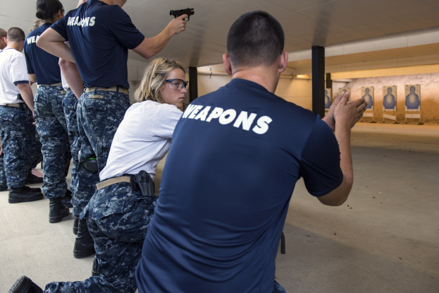 Eine Gruppe von Menschen in blauen Hemden und schwarzen Hosen steht in einem Schießstand, wobei eine Frau in der Mitte eine Waffe hält und Ziele an der Wand hinter ihnen sichtbar sind.