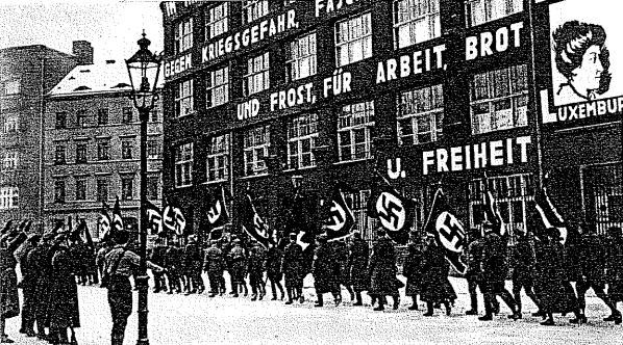 Schwarzes und weißes Bild einer Gruppe, die die Straße entlang marschiert und Fahnen und Plakate schwingt, mit einem Laternenmast auf der linken Seite und einer Tafel auf der rechten Seite mit der Aufschrift "Deutsche Nationalsozialistische Arbeiterpartei in Berlin".