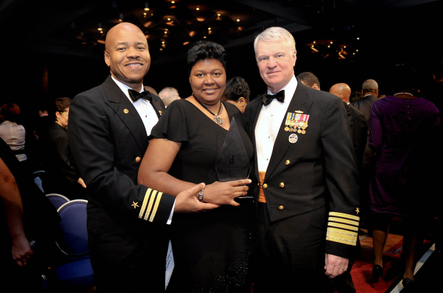 Zwei Männer in schwarzen Blazern und weißen Hemden stehen neben einer Frau in einem schwarzen Kleid, die eine Trophäe hält, auf einem roten Teppich, während andere sitzen und stehen in einem beleuchteten Innenraum.