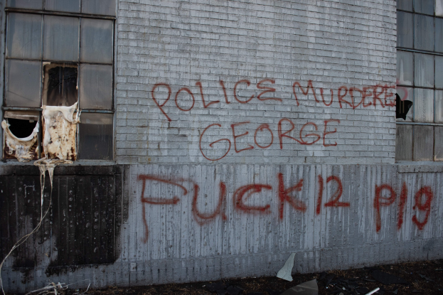 Graffiti auf einer Gebäudewand mit der Aufschrift 'Polizei Mord George Fuck 12 P19' mit Fenstern und verstreuten Gegenständen davor.