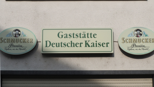 Gebäude mit drei wandmontierten Tafeln mit der Aufschrift "Gaststätte Deutscher Kaiser Schmucker" und einem Rollladen unten.