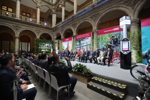 Eine Gruppe von Menschen sitzt auf Stühlen und schaut auf einen Mann auf einer Bühne, der ein Mikrofon hält, rechts daneben stehen Fahrräder und im Hintergrund sind Plakate, Pflanzen, Säulen, Geländer, Lampen, Fenster und eine Wand zu sehen.