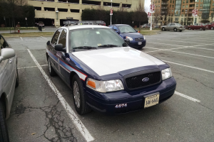 Polizeiauto auf einem Parkplatz mit einem Rad im Vordergrund, Bäume und Gebäude im Hintergrund unter einem klaren blauen Himmel.