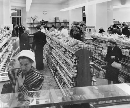 Schwarzes und weißes Foto von Menschen, die in einem Supermarkt einkaufen, mit Regalen voller Lebensmittel, einige halten Taschen, eine Uhr an der Wand und Deckenbeleuchtung.
