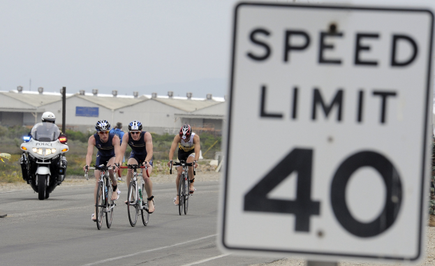 Gruppe von Radfahrern mit Helmen, die auf einer Straße mit einem Tempolimit-Schild fahren, mit Gebäuden und Bäumen im Hintergrund unter einem klaren blauen Himmel.
